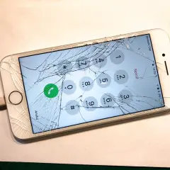 Zepsuty ekran telefonu z widocznymi pęknięciami, podłączony do ładowarki. Na ekranie widoczna klawiatura numeryczna i przycisk połączenia alarmowego.