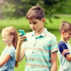 Dzieci w parku korzystają z telefonów. Chłopiec w centrum z niebieskim telefonem, dziewczynka po lewej z białym, a chłopiec po prawej z zielonym. Bezpieczne surfowanie dzięki Google VPN.
