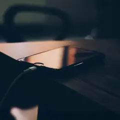 Telefon podłączony do ładowania, ale wciąż zastanawiasz się, dlaczego telefon wolno się ładuje? Ciemne tło i drewniany blat podkreślają problem.