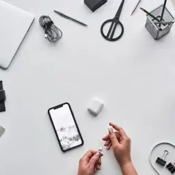 Ręce trzymające słuchawki AirPods, obok telefon z Androidem i etui. Czy airpods działają z androidem? Tak, można ich używać.