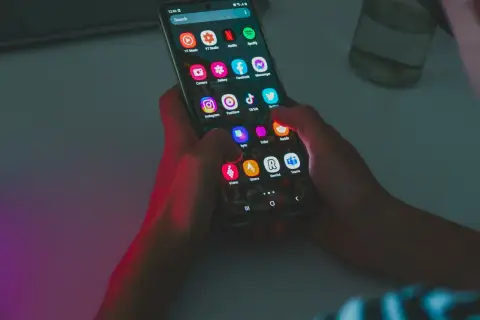 Dłonie trzymają smartfon z ikonami aplikacji. Widać Netflix, Spotify, Instagram, TikTok. Bateria szybko się rozładowuje, co widać po intensywnym użytkowaniu.