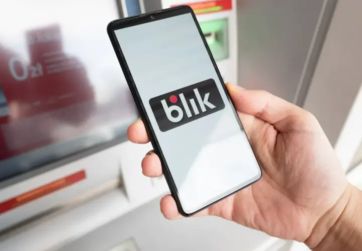 Dłoń trzyma smartfon z logo Blik przed bankomatem. Uważaj na oszustów, którzy mogą próbować wyłudzić dane.