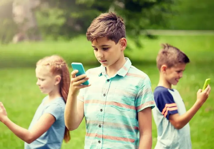 Dzieci w parku korzystają z telefonów. Chłopiec w centrum z niebieskim telefonem, dziewczynka po lewej z białym, a chłopiec po prawej z zielonym. Bezpieczne surfowanie dzięki Google VPN.