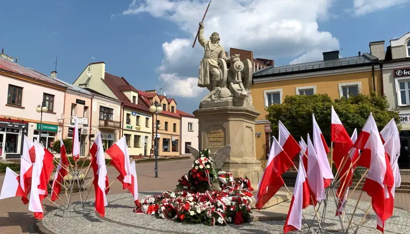 Pomnik z polskimi flagami, w tym kierunkowy 15, na placu przed kolorowymi kamienicami.