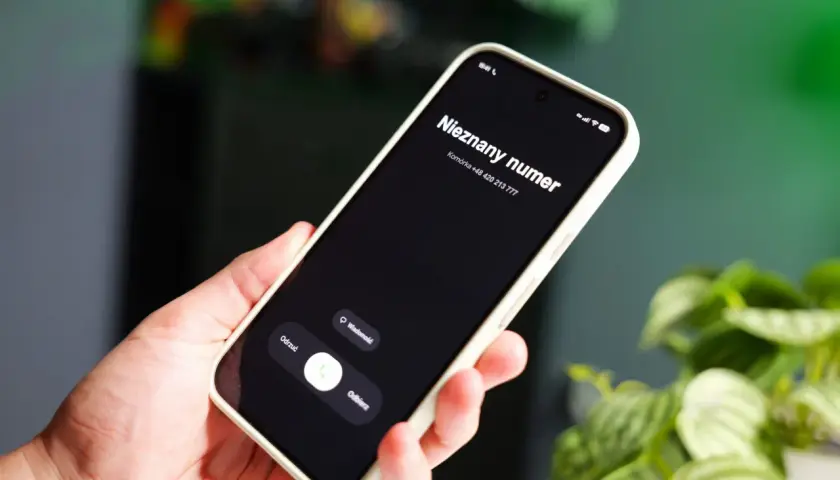 Dłoń trzyma smartfon z ekranem blokady, na którym widnieje "Nieznany numer" i numer telefonu. W tle zielone rośliny.