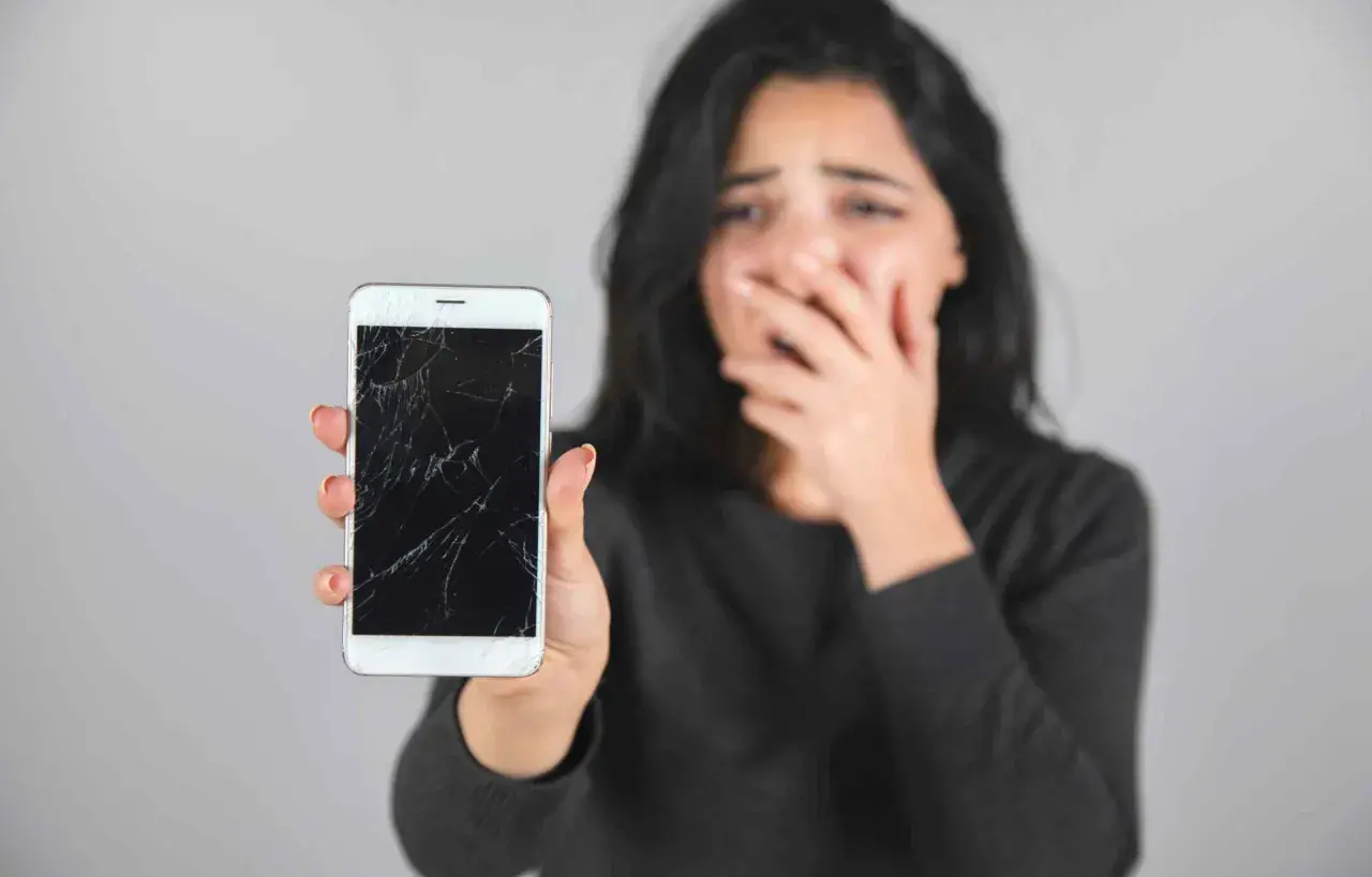 Zrozpaczona kobieta pokazuje pęknięty, czarny ekran w telefonie.