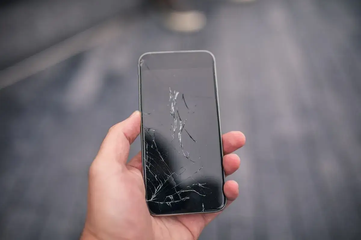 Dłoń trzyma telefon z pękniętym, zepsutym ekranem. Sieć pęknięć przecina czarną powierzchnię wyświetlacza.